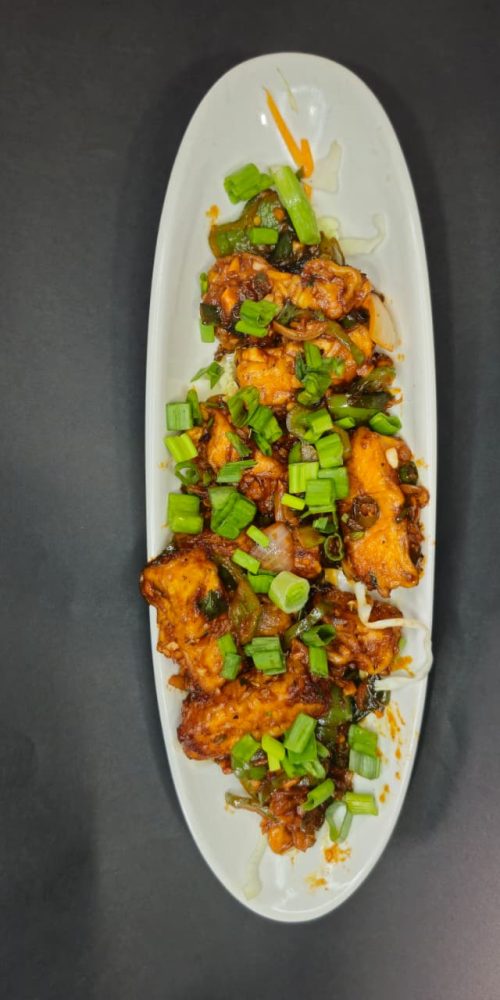 Top-down view of a spicy Indo-Chinese chilli paneer starter garnished with green spring onions on a long white oval platter against a black background.